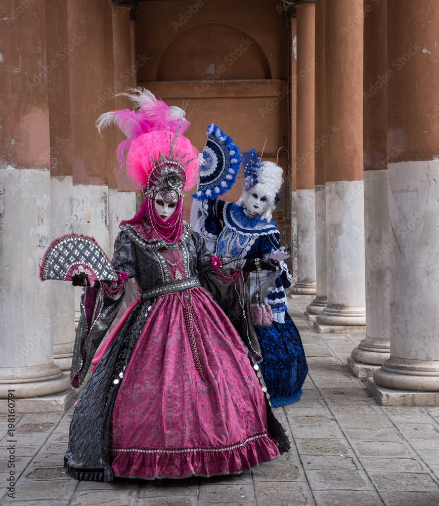Two women in masks and ornate blue and pink costumes standing in front of pillars at a monastery during Venice Carnival (Carnival di Venezia).