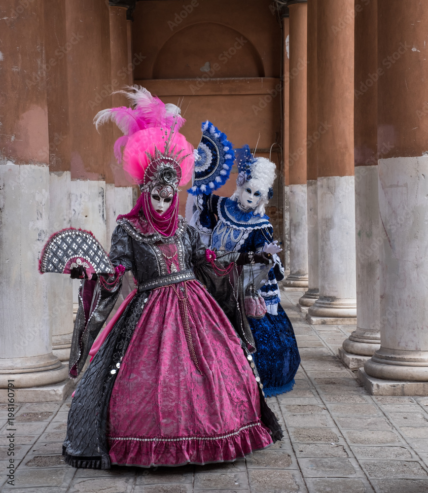 Two women in masks and ornate blue and pink costumes standing in front of pillars at a monastery during Venice Carnival (Carnival di Venezia). Two women in masks and ornate blue and pink costumes standing in front of pillars at a monastery during Venice Carnival (Carnival di Venezia).