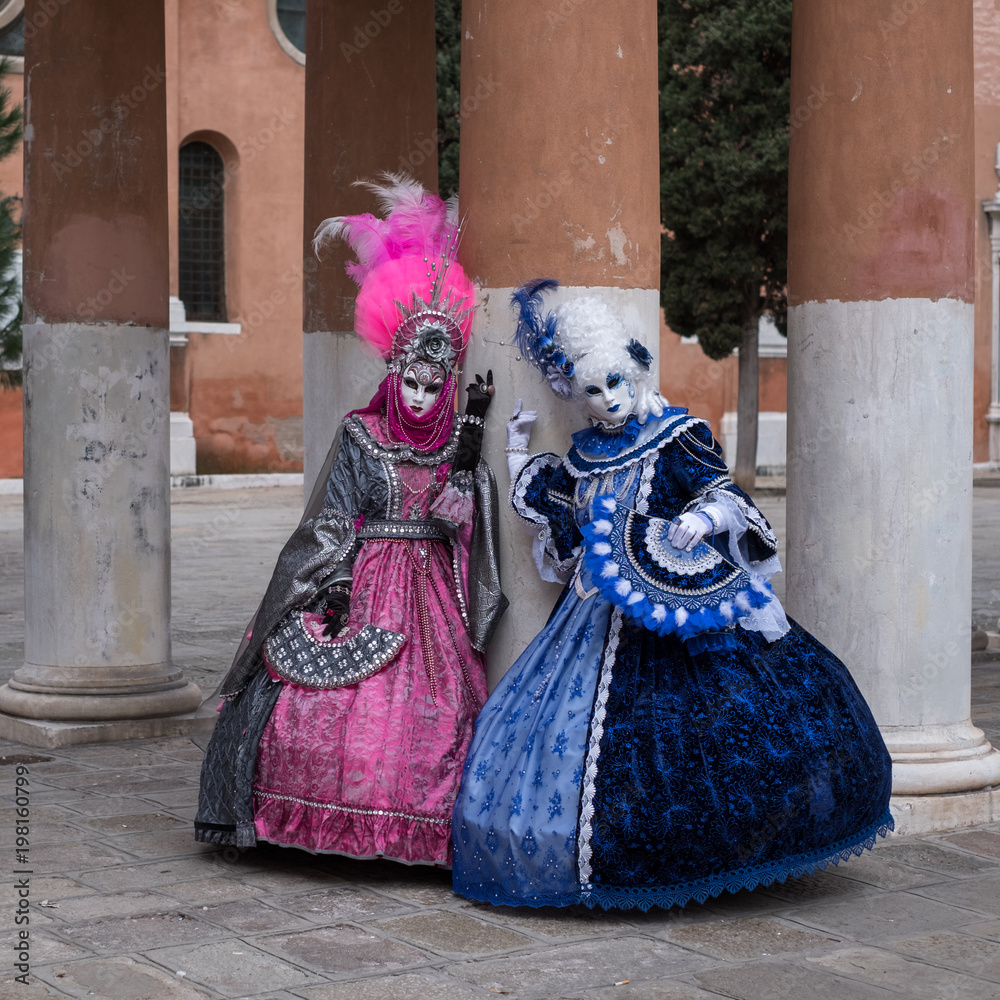 Two women in masks and ornate blue and pink costumes standing in front of pillars at a monastery during Venice Carnival (Carnival di Venezia). Two women in masks and ornate blue and pink costumes standing in front of pillars at a monastery during Venice Carnival (Carnival di Venezia).