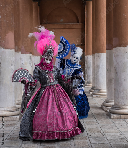 Two women in masks and ornate blue and pink costumes standing in front of pillars at a monastery during Venice Carnival (Carnival di Venezia). © Lois GoBe
