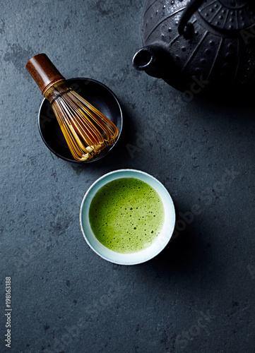 Cup of Matcha Green Tea on dark stone background; tea whisk, cast ron tea pot, japanese ceramic