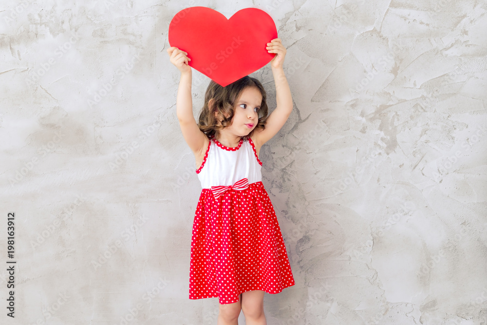 Happy funny baby girl with red balloon in the shape of a heart near the empty grey concrete walls. Concept of the celebration. Happy funny baby girl with red balloon in the shape of a heart near the empty grey concrete walls. Concept of the celebration.