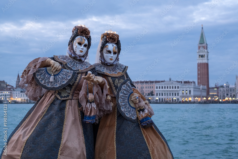 Venice Italy, February 2018. Two women in traditional costumes and masks, with decorated fans, standing in front of the Grand Canal with San Marco in the background, during the Venice Carnival