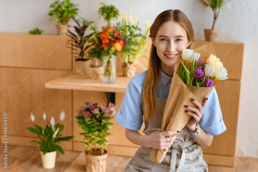 beautiful young florist holding bouquet of tulips and smiling at camera in flower shop beautiful young florist holding bouquet of tulips and smiling at camera in flower shop