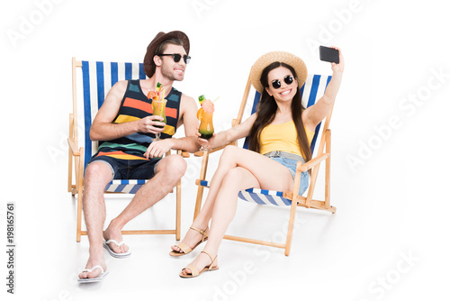 couple relaxing on beach chairs with cocktails and taking selfie, isolated on white