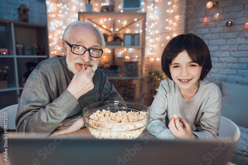 Grandfather and grandson are watching movie with popcorn at night at home.