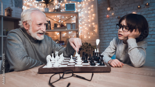 Grandfather and grandson are playing chess together at night at home. Granddad is winning.