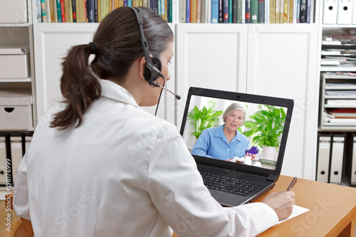 Doctor of geriatrics in her surgery office with headset in front of her laptop during a video call with a senior patient about her prescribed drugs. photo