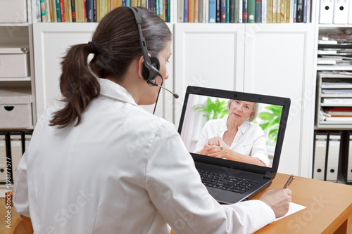 Fotografija Doctor with headset and laptop, taking notes during an online call with a patient showing a large mole, teledermatology concept