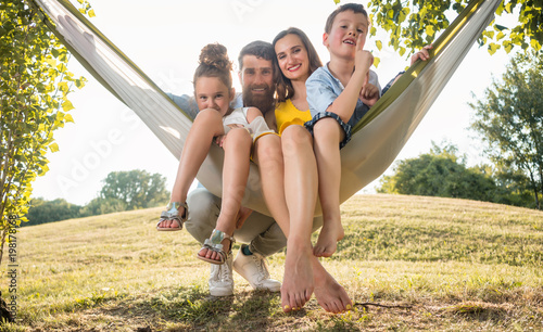 Family portrait with a beautiful mother of two playful children swinging in a hammock while looking at camera next to her husband outdoors in summer 
