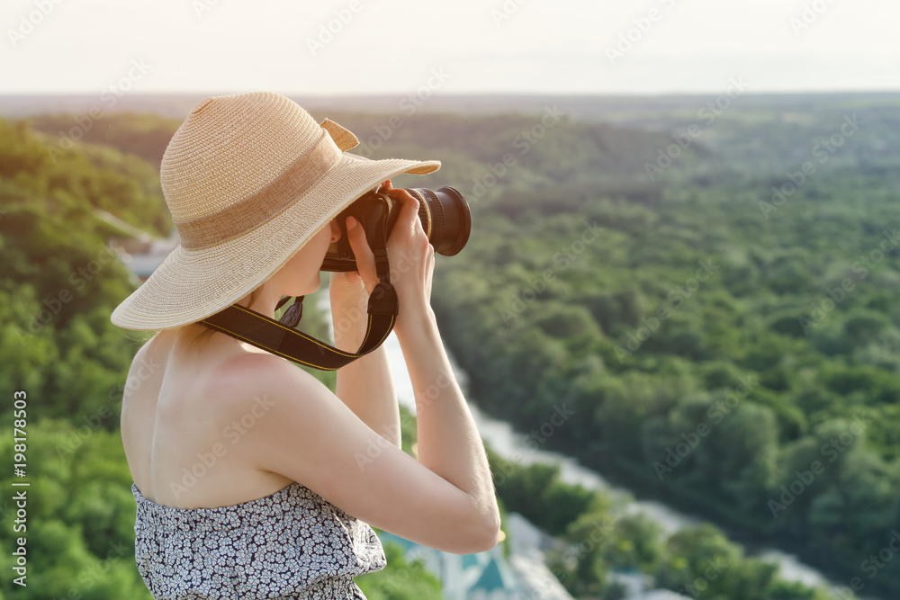 Girl sits on hill and takes pictures against the background of a forest and river
