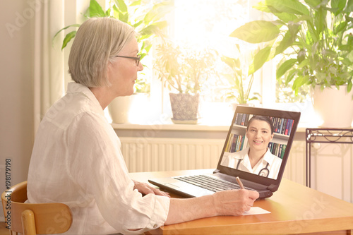 Teleconsultation concept: senior woman in her sunny living room in front of a laptop taking notes during a video call with her doctor. photo