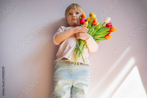 Powazna little girl o blond wlosach trzyma bukiet kolorowych tulipanow w rekach w slonecznym pokoju w domu. Concept swieta. photo