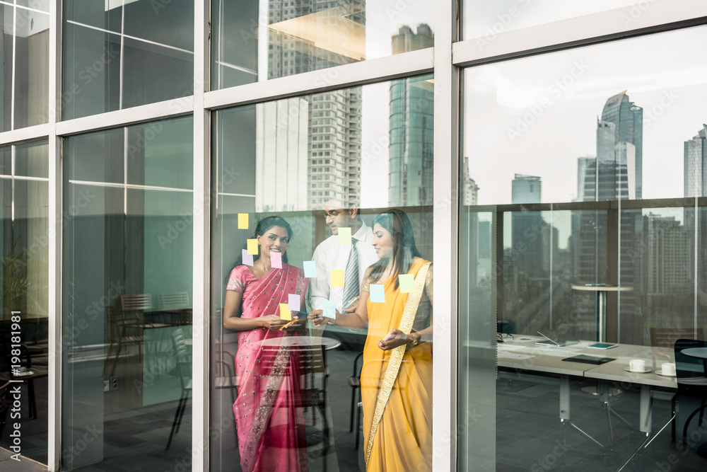 Three Indian employees sticking reminders on glass wall with business tasks and deadlines in the office