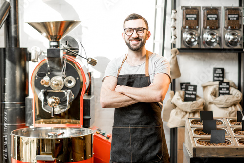 Portrait of a handsome barista in uniform standing in the coffee shop with roaster machine on the background