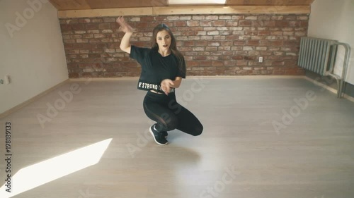 Beautiful athletic girl in black dancing booty dance twerk in studio sitting on floor on brown background. Vogue or hip hop and jumping. Long shot.