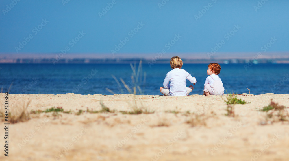 two young kids, brothers sitting on sandy beach in the morning near the lake