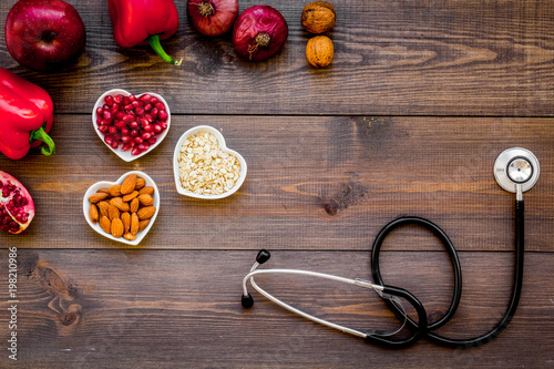 Products good for heart and blood vessels. Vegetables, fruits, nuts in heart shaped bowl near stethoscope on dark wooden background top view copy space
