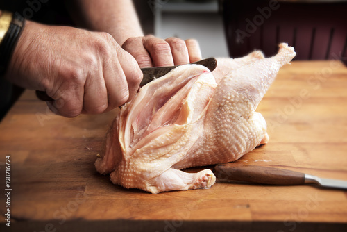 Men's hands prepare a raw chicken with a kitchen knife for cooking on a rustic wooden butcher block