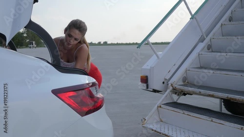 rich girl near a private jet pulls out travel bag from trunk of avtomobile at airport and rises at staircase in slow motion photo