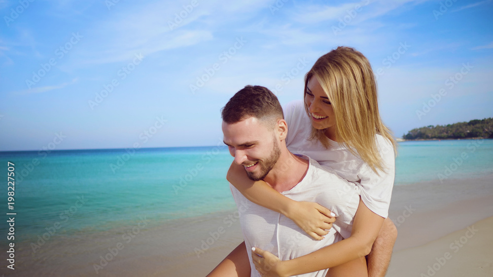 Beautiful happy couple having fun, doing piggyback on tropical sandy beach over sea and sky background