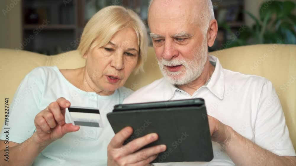 Cheerful good-looking senior couple sitting on couch at home using tablet. Buying online with credit card on computer. Consumerism concept. Active modern life after retirement. Cheerful good-looking senior couple sitting on couch at home using tablet. Buying online with credit card on computer. Consumerism concept. Active modern life after retirement.