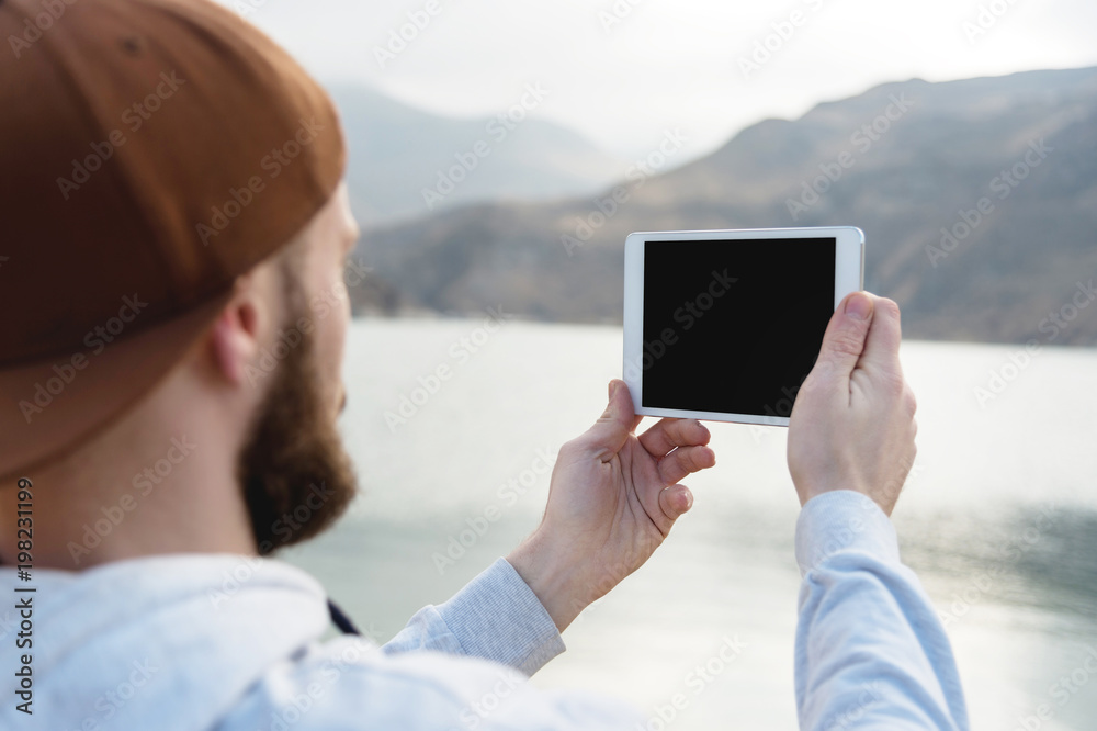 Hipster person holding in hands digital tablet with empty blank screen, man photograph on computer on background nature outdoor landscape mock up technology blur male hands tourist using gadget