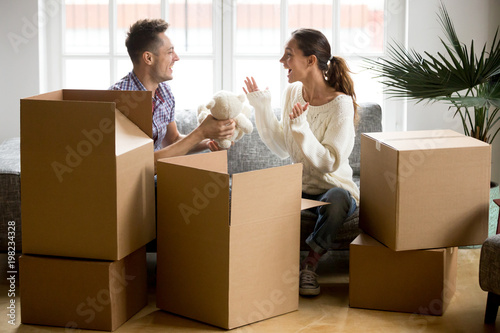 Young happy couple having fun packing boxes on moving day, loving boyfriend making gift holding cute toy found in belongings giving white teddy bear to excited girlfriend while unpacking in new home