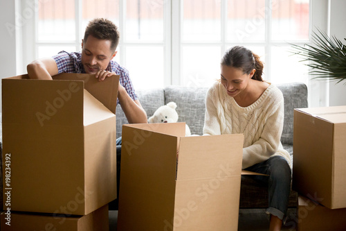 Young smiling couple unpacking cardboard boxes together sitting on sofa in modern living room, moving out or settle in new home concept, man and woman packing belongings stuff preparing to relocate