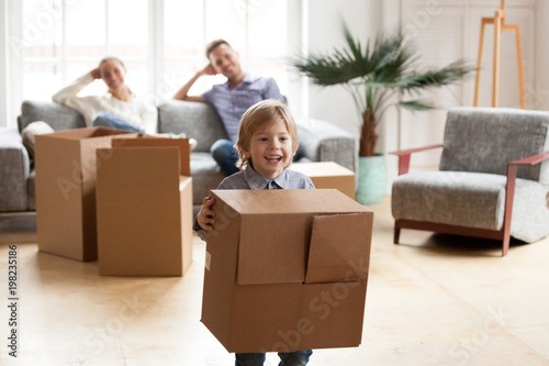 Excited little boy playing with cardboard box on moving day, parents relaxing on sofa while their happy active son enjoying packing relocating into new home, cute kid having fun in modern living room