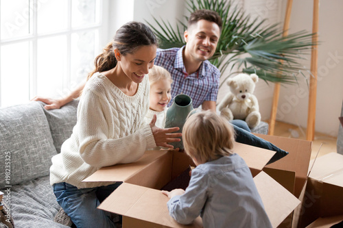 Happy family with two kids unpacking boxes after relocation moving into or settling in new home concept, excited small children helping parents with belongings sitting on sofa in living room together photo