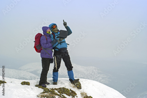 two hikers on a winter mountain peak with a laptop