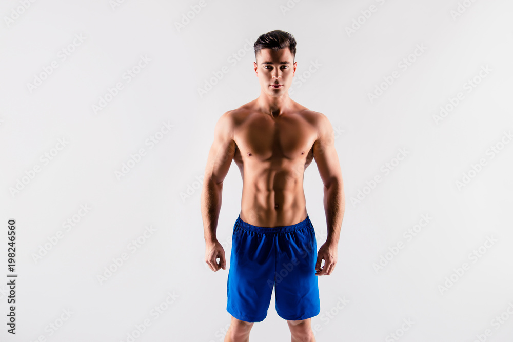 Portrait of handsome serious confident concentrated muscular sportsman clothed in blue loose shorts, he is demonstrating his perfect ideal body constitution, isolated on grey background, copyspace