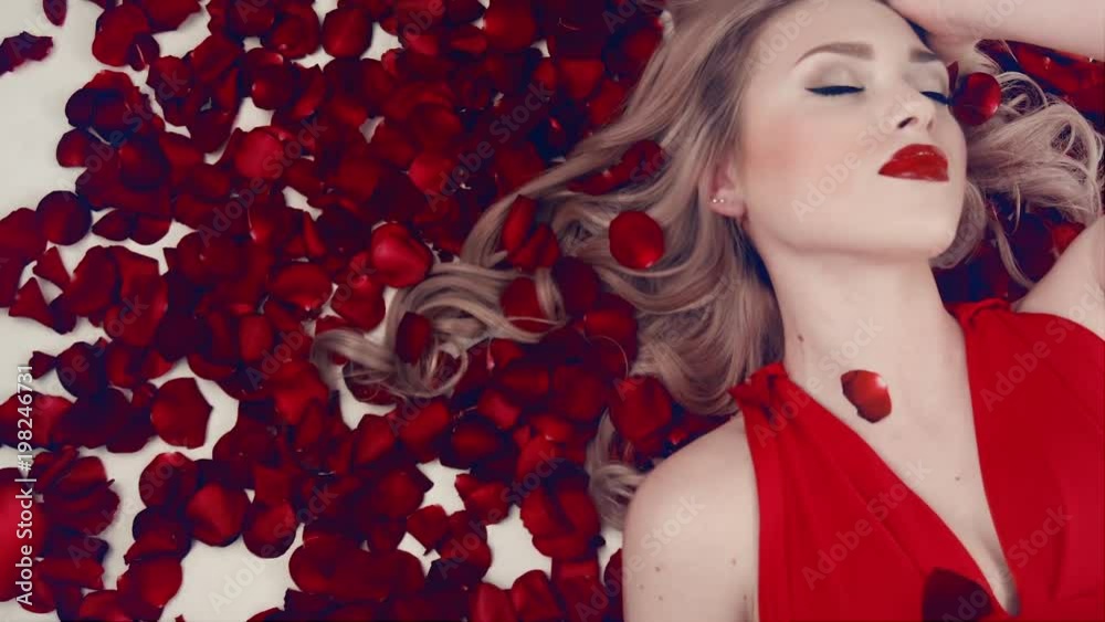 Beautiful blond girl with red lips and in a red dress lies in the petals of red roses. Rose petals fall on the girl who lies in the petals. Slow motion. Valentine's Day. Beauty is American. Beautiful blond girl with red lips and in a red dress lies in the petals of red roses. Rose petals fall on the girl who lies in the petals. Slow motion. Valentine's Day. Beauty is American.