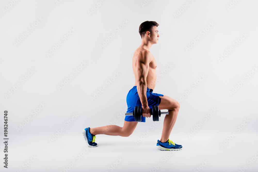 Side profile view portrait of sportive handsome  muscular shirtless, wearing blue shorts and sneakers bodybuilder, he is holding dumbbells and doing sit-ups, isolated on grey background, copy-space