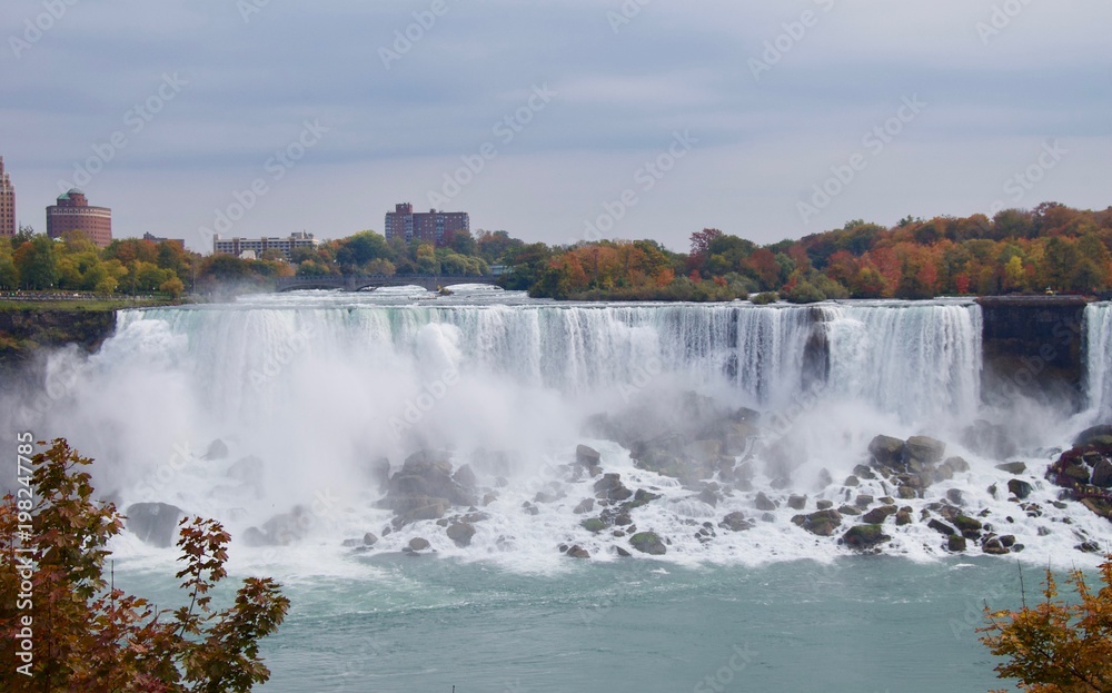 Beautiful and impressive panorama of the Niagara Falls in Ontario (Canada) on a bright autumn day with water crashing down the falls onto rocks creating lots of mist in front of the city skyline Beautiful and impressive panorama of the Niagara Falls in Ontario (Canada) on a bright autumn day with water crashing down the falls onto rocks creating lots of mist in front of the city skyline