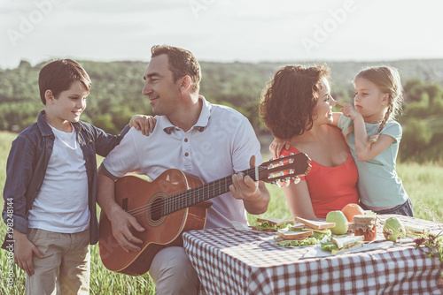 Joyful father and son are singing favorite song to guitar. Woman is playing with daughter and smiling. They are sitting at table on summer meadow