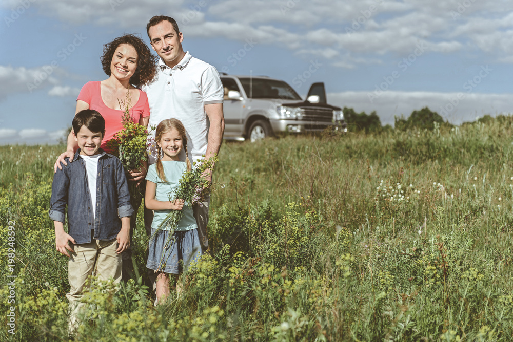 Fototapeta premium Happy together. Full length portrait of cheerful father and mother spending time with their kids on grassland. Girl is holding bouquet of wild flowers while boy is smiling. Copy space