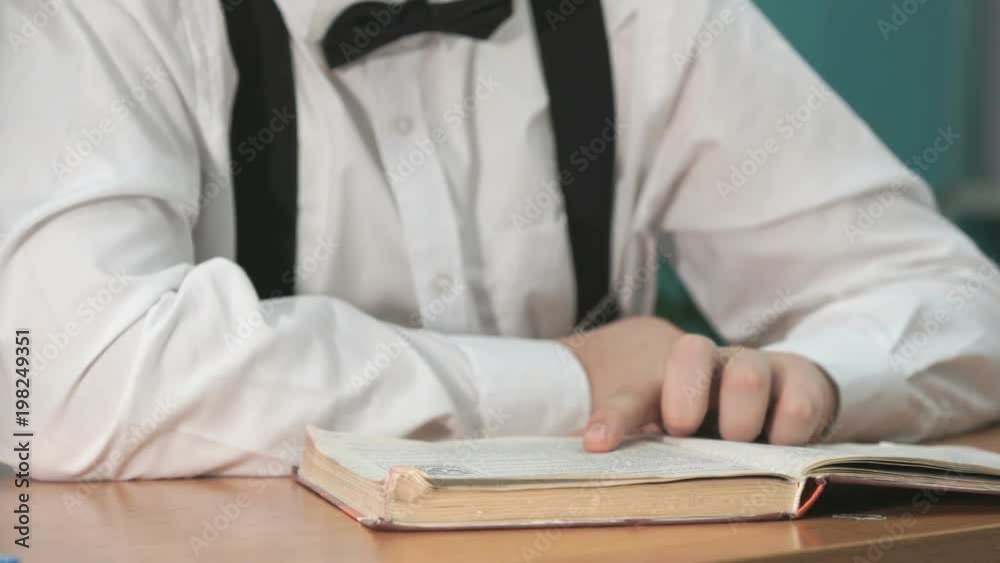 Unknown student boy dressed in white shirt with bow tie sitting at a desk at the university and reading a book. Close-up