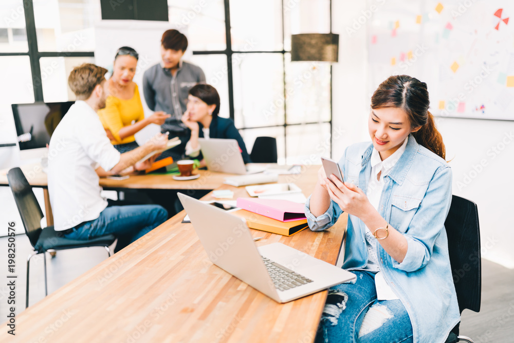 Obraz premium Asian woman using smart phone app check email or text message, multiethnic diverse team discussing in background. Business cooperate teamwork, strategy brainstorm meeting, or SME ecommerce technology