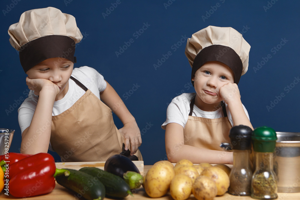 Portrait of two little boys dressed in chef uniform having bored looks, leaning on their elbows, don't want to help mother cooking dinner, standing at kitchen table with fresh organic vegetables Portrait of two little boys dressed in chef uniform having bored looks, leaning on their elbows, don't want to help mother cooking dinner, standing at kitchen table with fresh organic vegetables