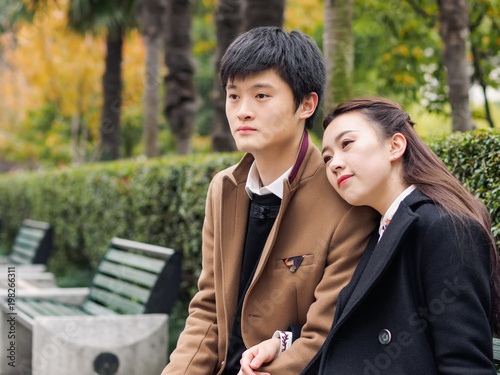 Portrait of couple of Chinese young people sit in bench in autumn park, hand in hand and hug together, lover concept.