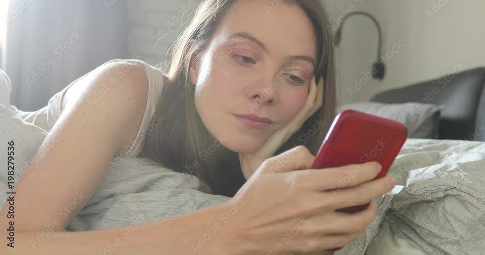 Happy woman lying on bed smiling while reading a text message from someone Happy woman lying on bed smiling while reading a text message from someone