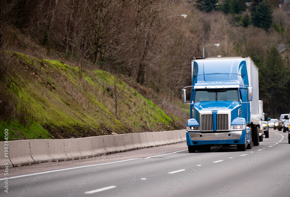 Modern blue big rig semi truck with semi trailer going on the road in front of another traffic transport