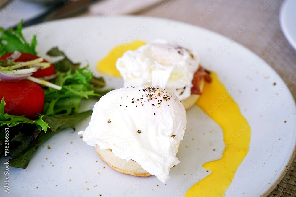 Two eggs benedicts, breakfast menu, consist of eggs, english muffin, canadian bacon and Hollandaise sauce served with salad placed on white dish. Two eggs benedicts, breakfast menu, consist of eggs, english muffin, canadian bacon and Hollandaise sauce served with salad placed on white dish.