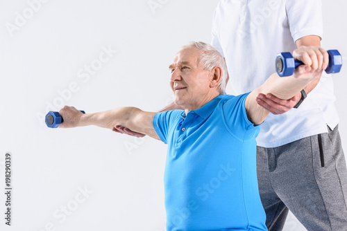 partial view of rehabilitation therapist assisting senior man exercising with dumbbells on grey background