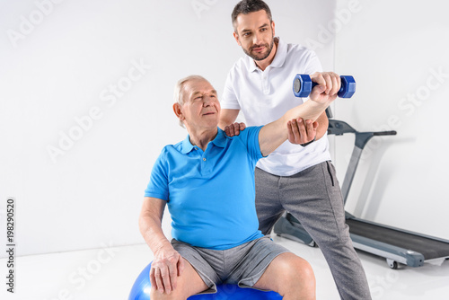 portrait of rehabilitation therapist assisting senior man exercising with dumbbell on grey backdrop
