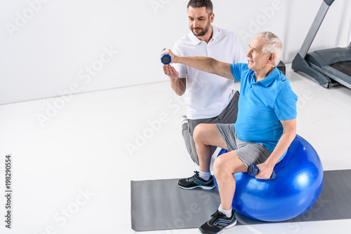 high angle view of rehabilitation therapist assisting senior man exercising with dumbbells on fitness ball