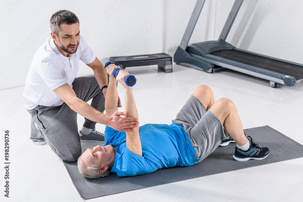 rehabilitation therapist assisting senior man exercising with dumbbells on mat