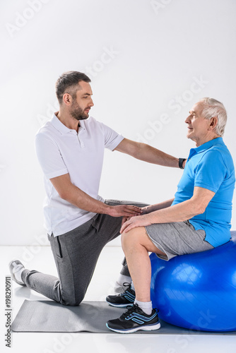 side view of rehabilitation therapist assisting senior man exercising on fitness ball on grey backdrop